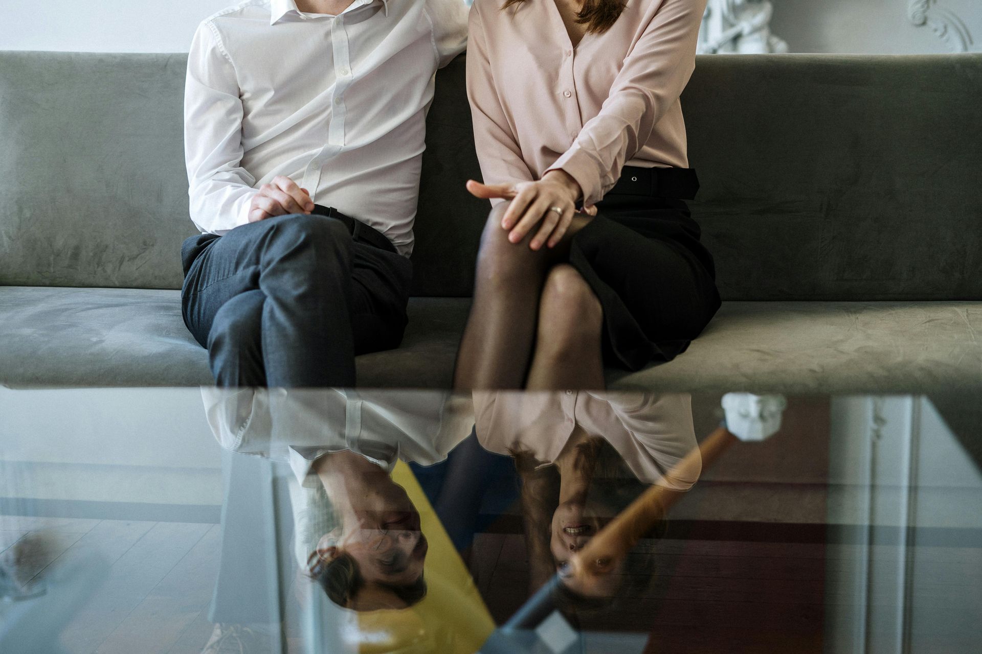 A couple sits on a couch; reflected in a glass table below. The man has an arm around the woman's shoulder.