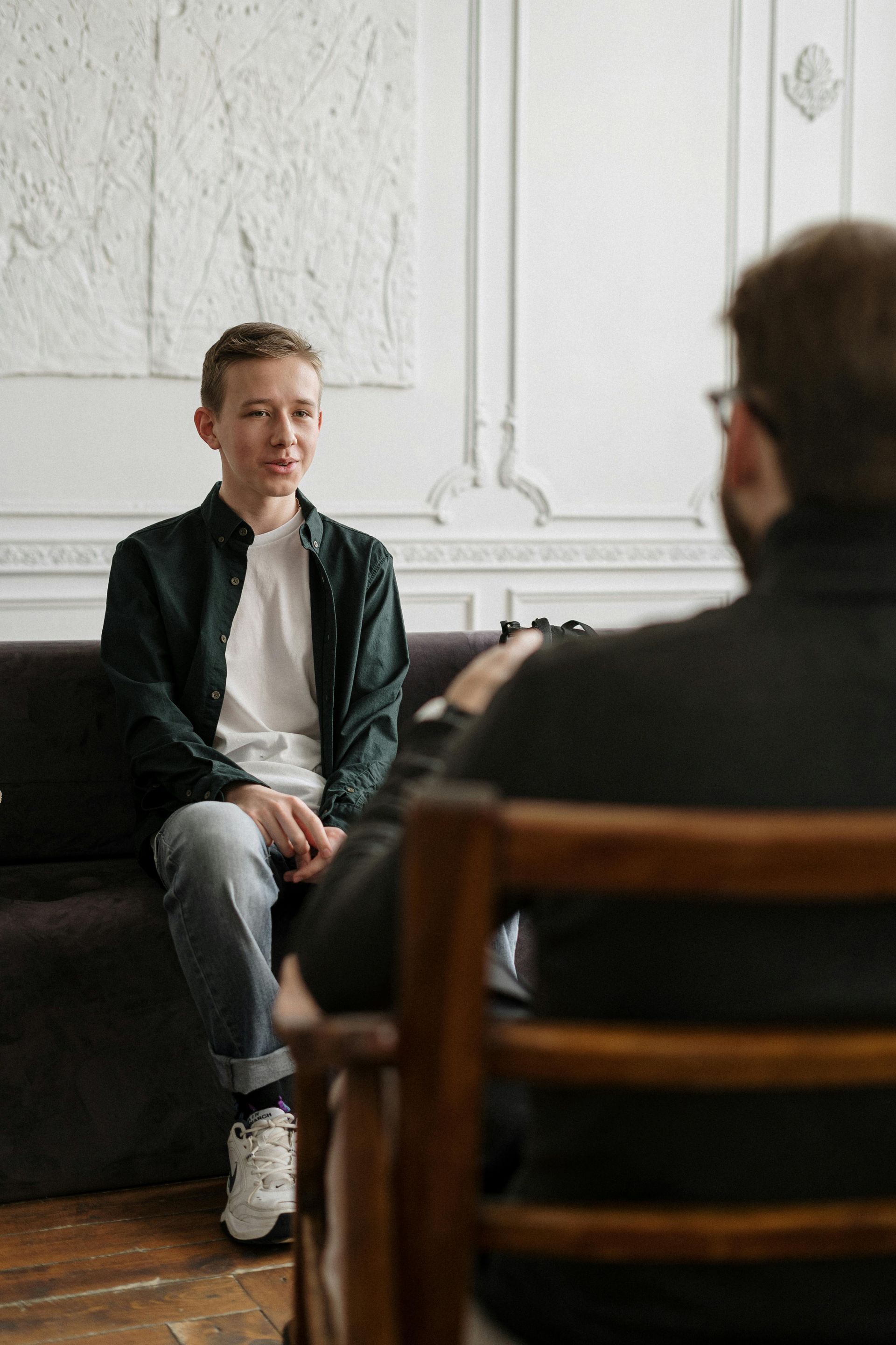 A person sits on a couch, talking to a person in a chair. Interior setting.