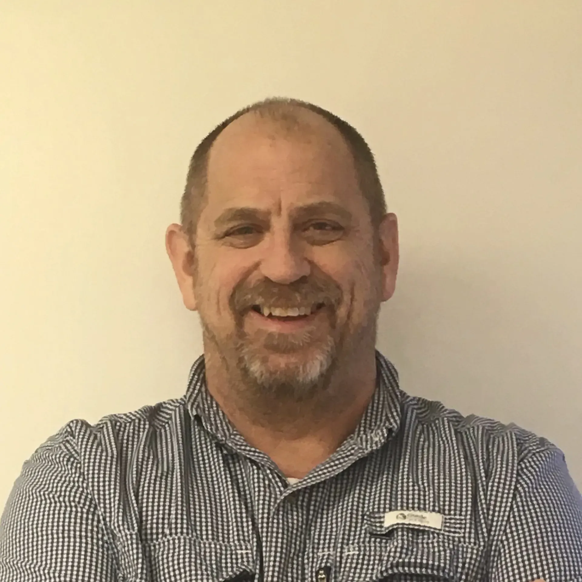 A smiling man with a beard, wearing a blue and white checkered short-sleeve button-down shirt against a plain wall.