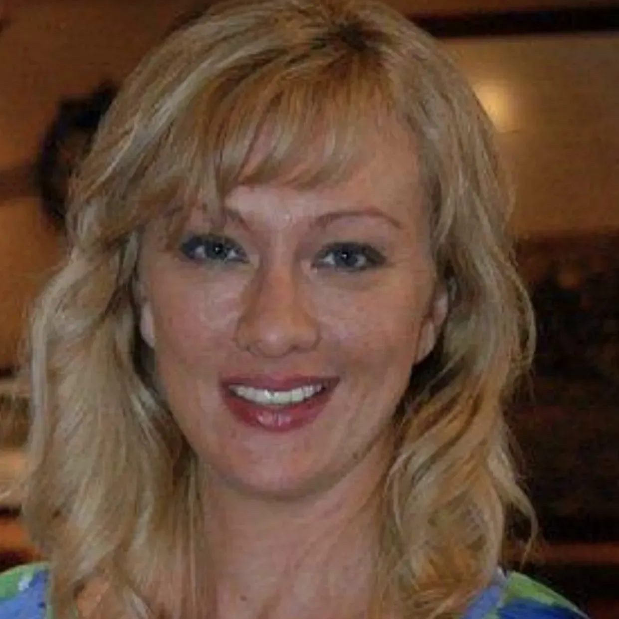 A smiling person with shoulder-length blonde hair, wearing a patterned top in a softly lit indoor setting.