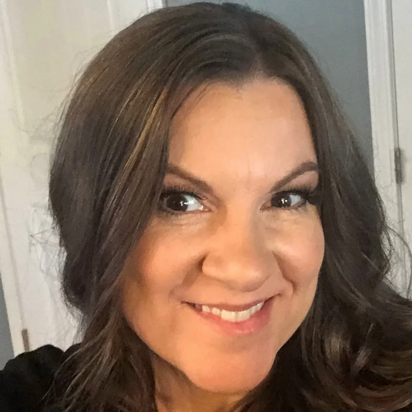 A headshot of a person with long, dark, wavy hair, smiling at the camera with a neutral, soft indoor background.