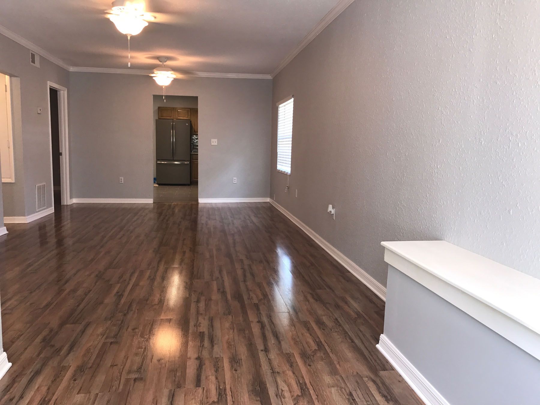 Empty living room with gray walls, wood-look flooring, two ceiling light fixtures, and a view into a kitchen.