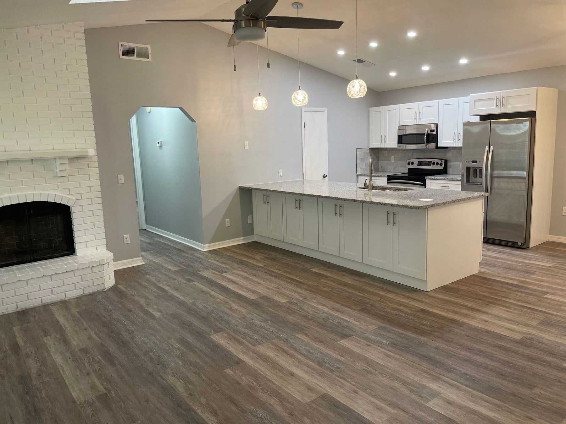 A bright open-concept kitchen and living area featuring a white brick fireplace, wood floors, and a white kitchen island.