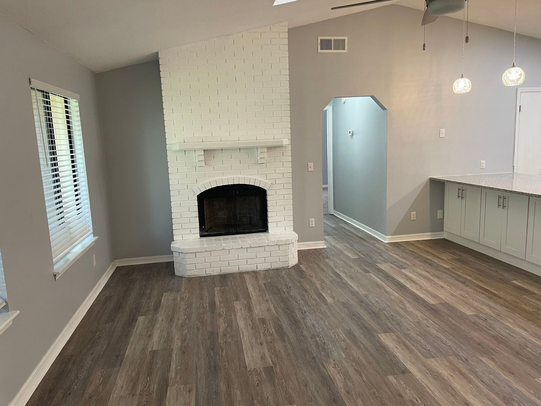 A bright room with grey walls, white-painted brick fireplace, wood-look flooring, and a kitchen counter on the right.