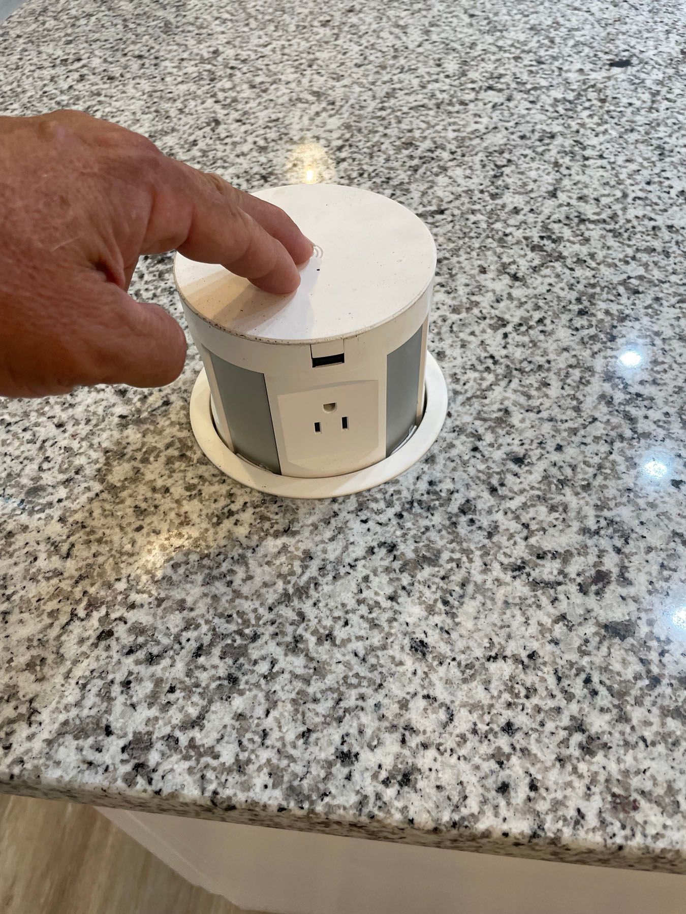 A hand presses down on a white, retractable pop-up electrical outlet installed in a speckled granite countertop.