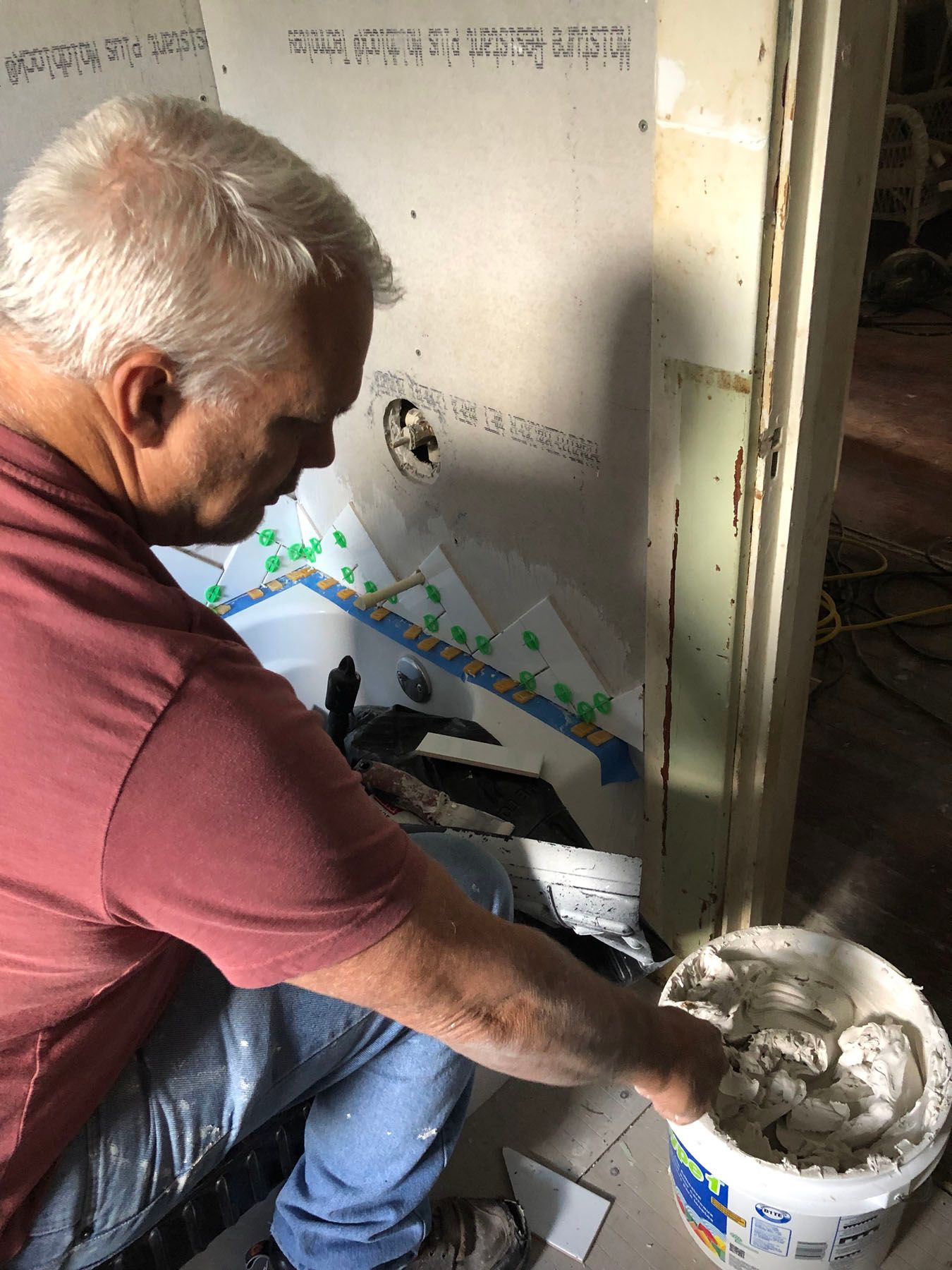 A person in a maroon shirt reaches into a bucket of white mortar while installing tiles on a bathroom wall.
