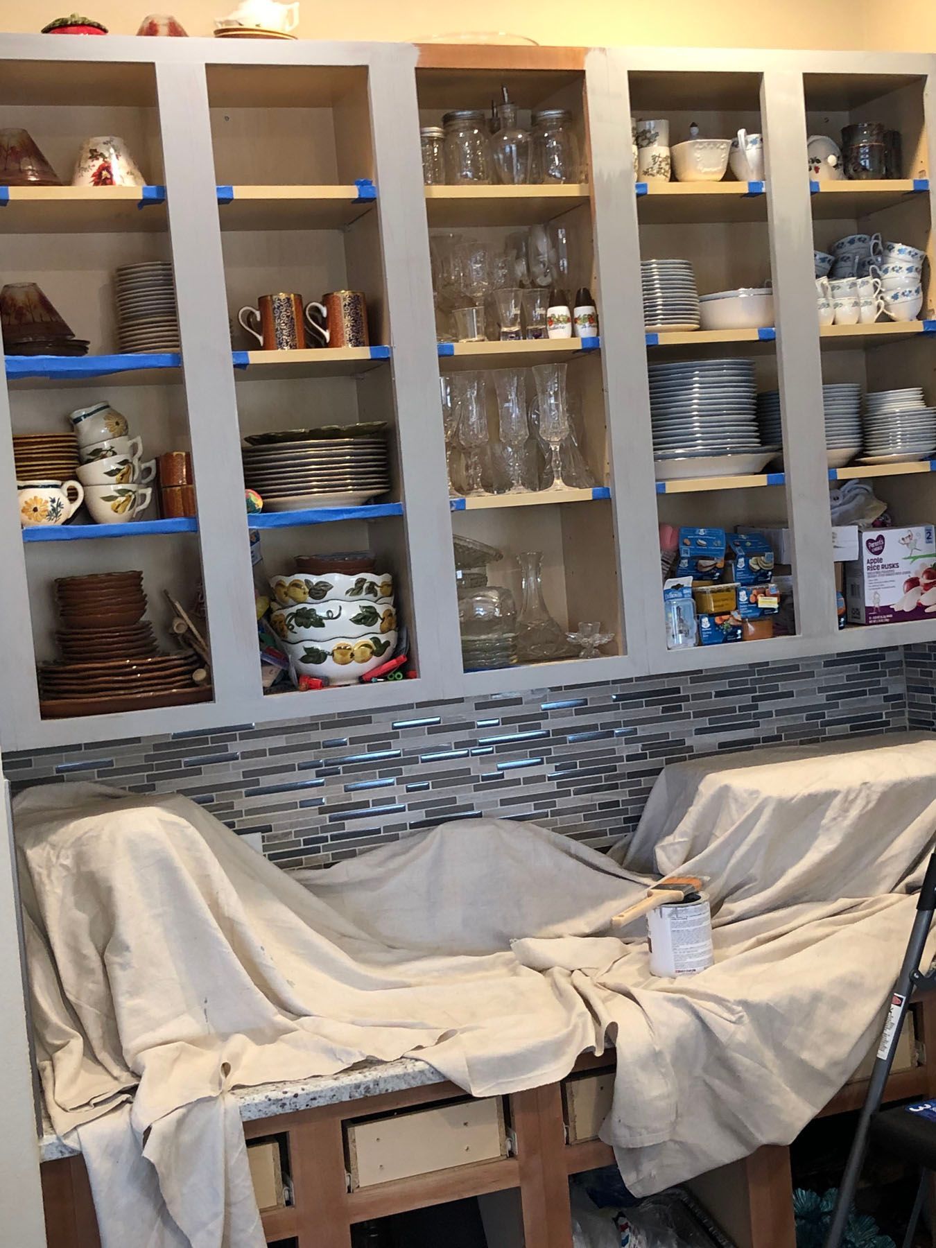 A kitchen with shelves containing dishes, currently undergoing cabinet painting with blue tape on edges and drop cloths.