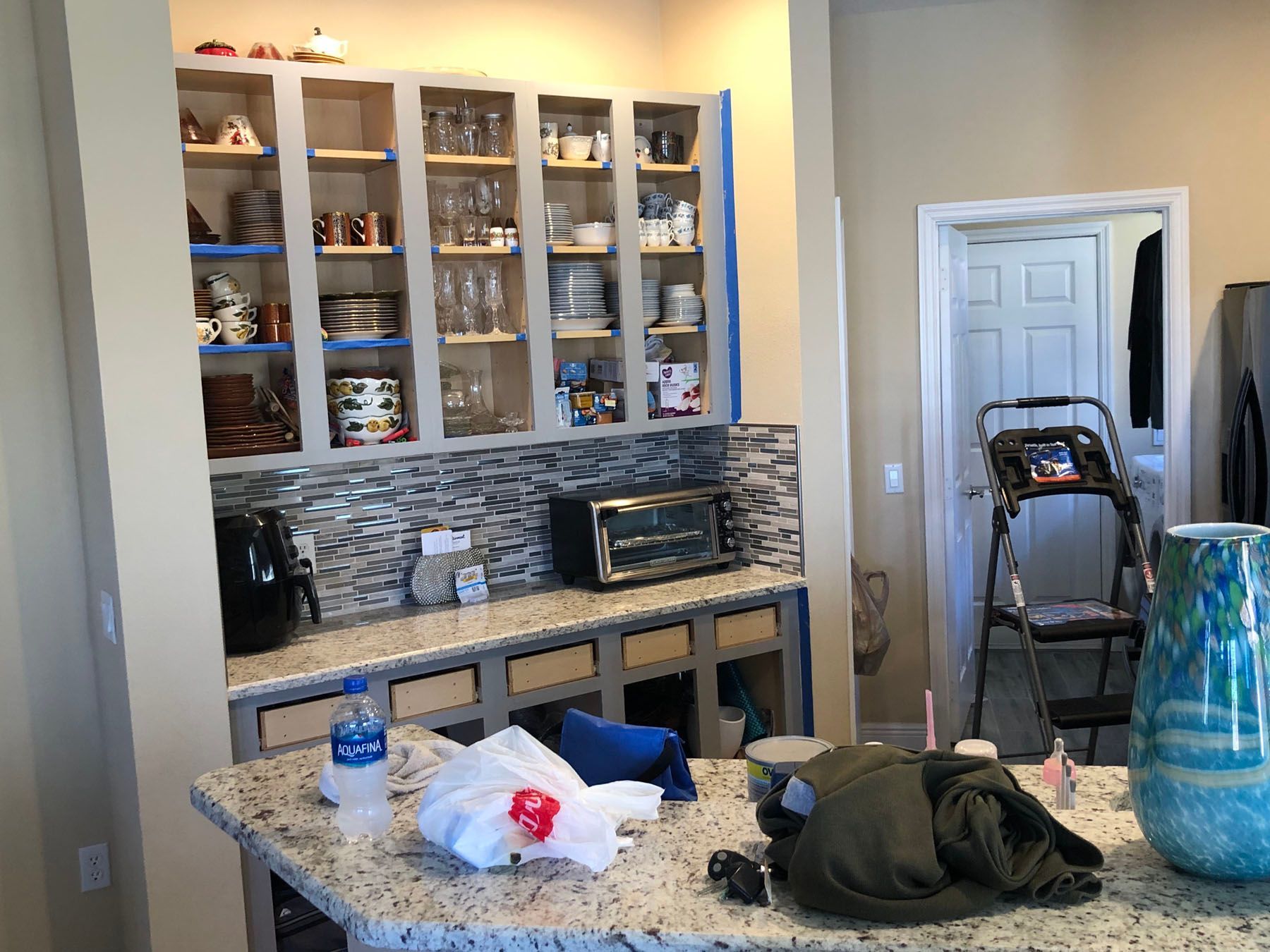 A kitchen undergoing renovations features open cabinetry, a tiled backsplash, and granite countertops with scattered items.