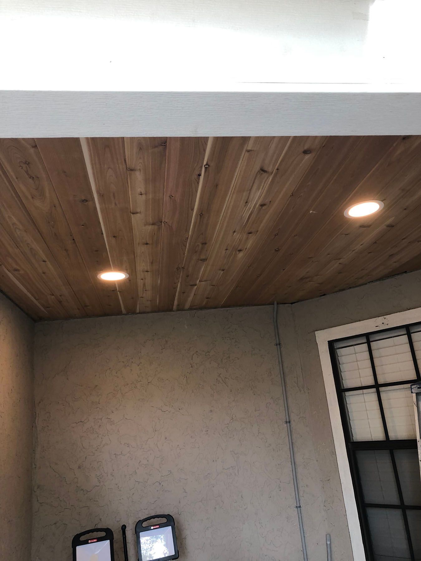 A wooden tongue-and-groove ceiling with two recessed lights above a textured gray wall and a window with white trim.