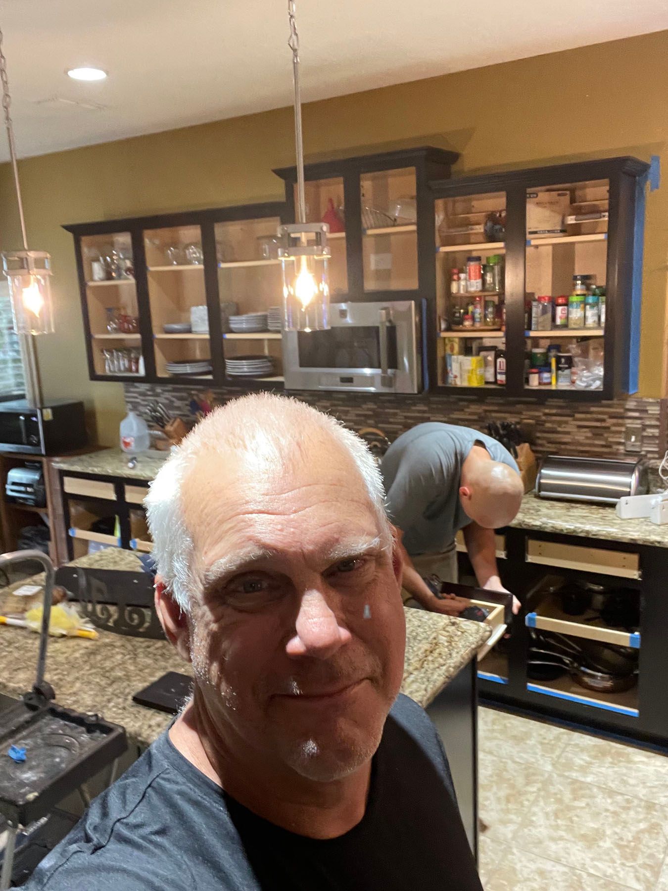 A person smiling in the foreground of a kitchen during a home renovation, with another person working on cabinets behind.