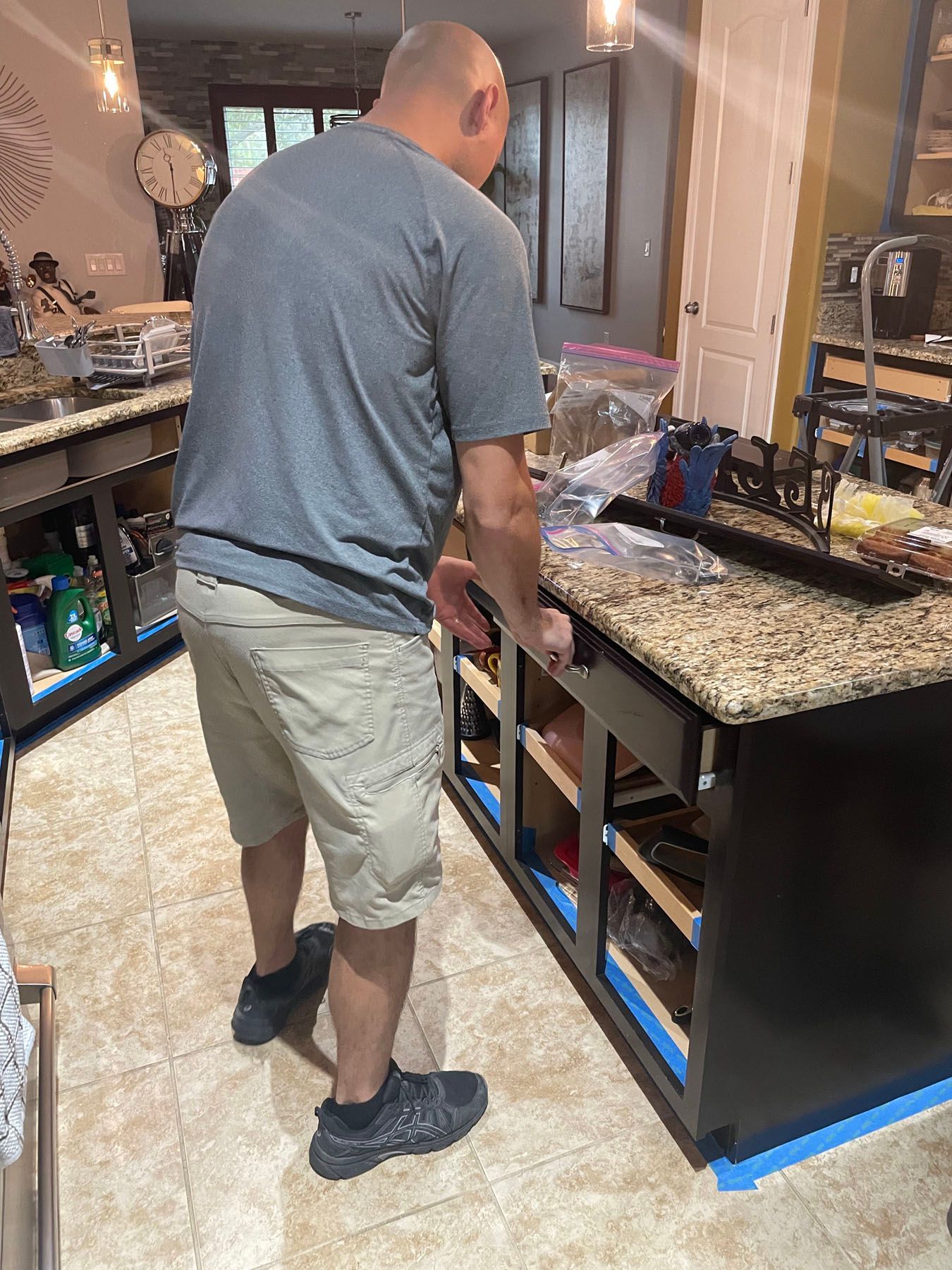 A person wearing a grey shirt and khaki shorts works on an open drawer of a kitchen island with granite countertops.