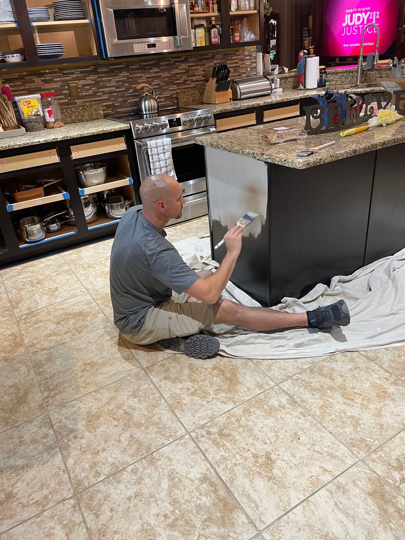A person sits on a drop cloth in a kitchen, painting the side of a black kitchen island with a white paintbrush.