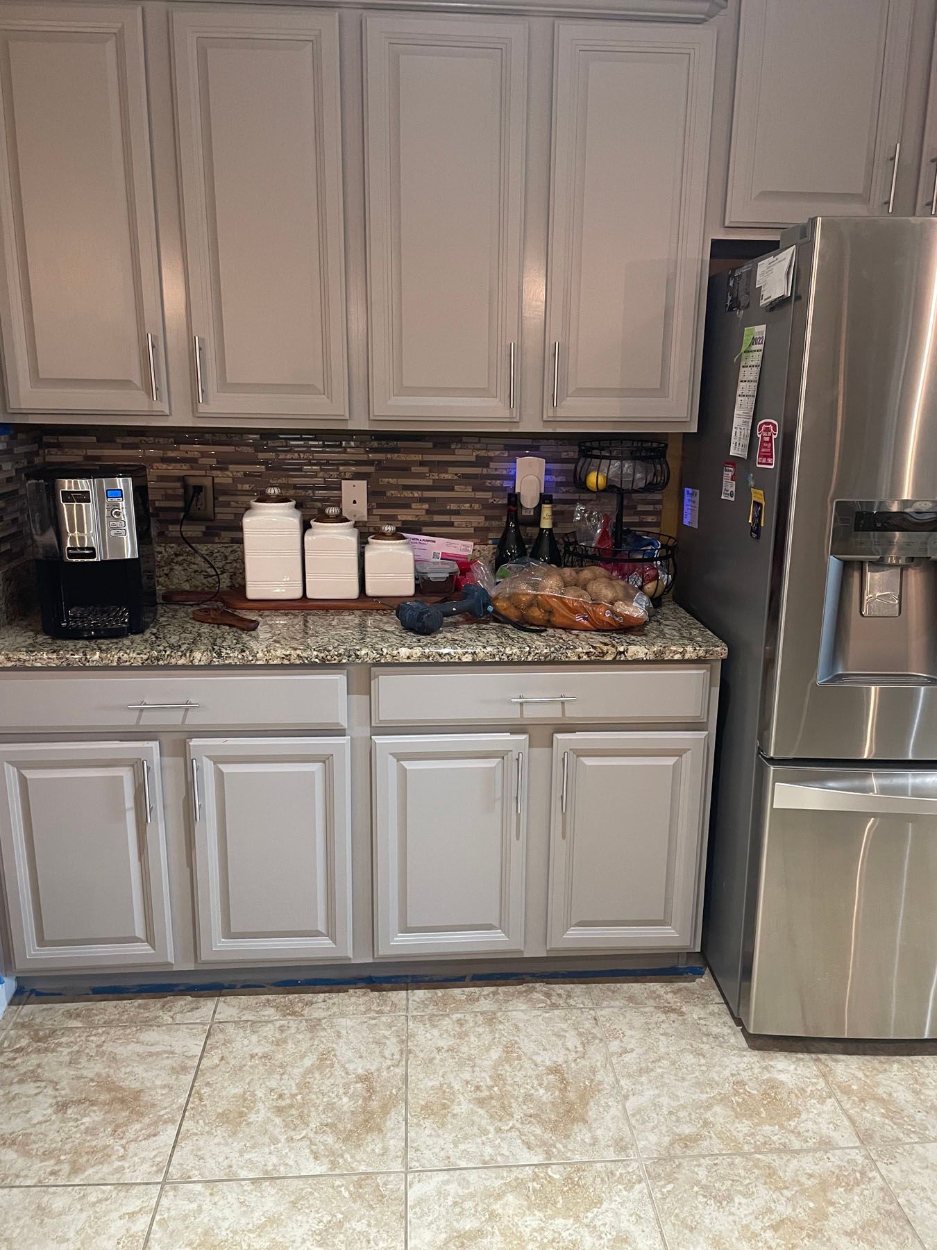 Gray kitchen cabinets with granite countertops, a coffee maker, kitchen canisters, and a stainless steel refrigerator.