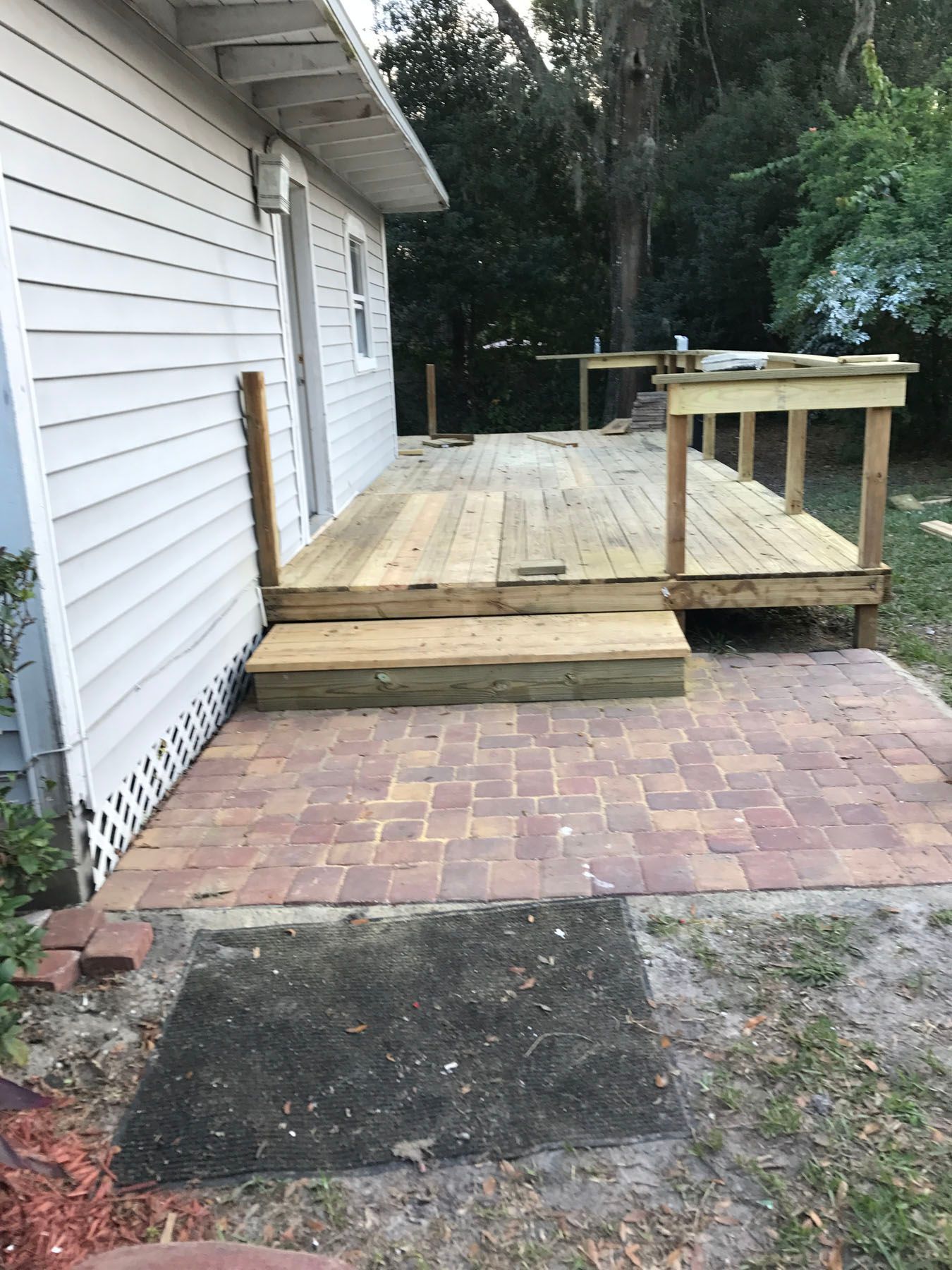 A side view of a house exterior featuring a newly built wooden deck with stairs leading down to a paved brick patio.