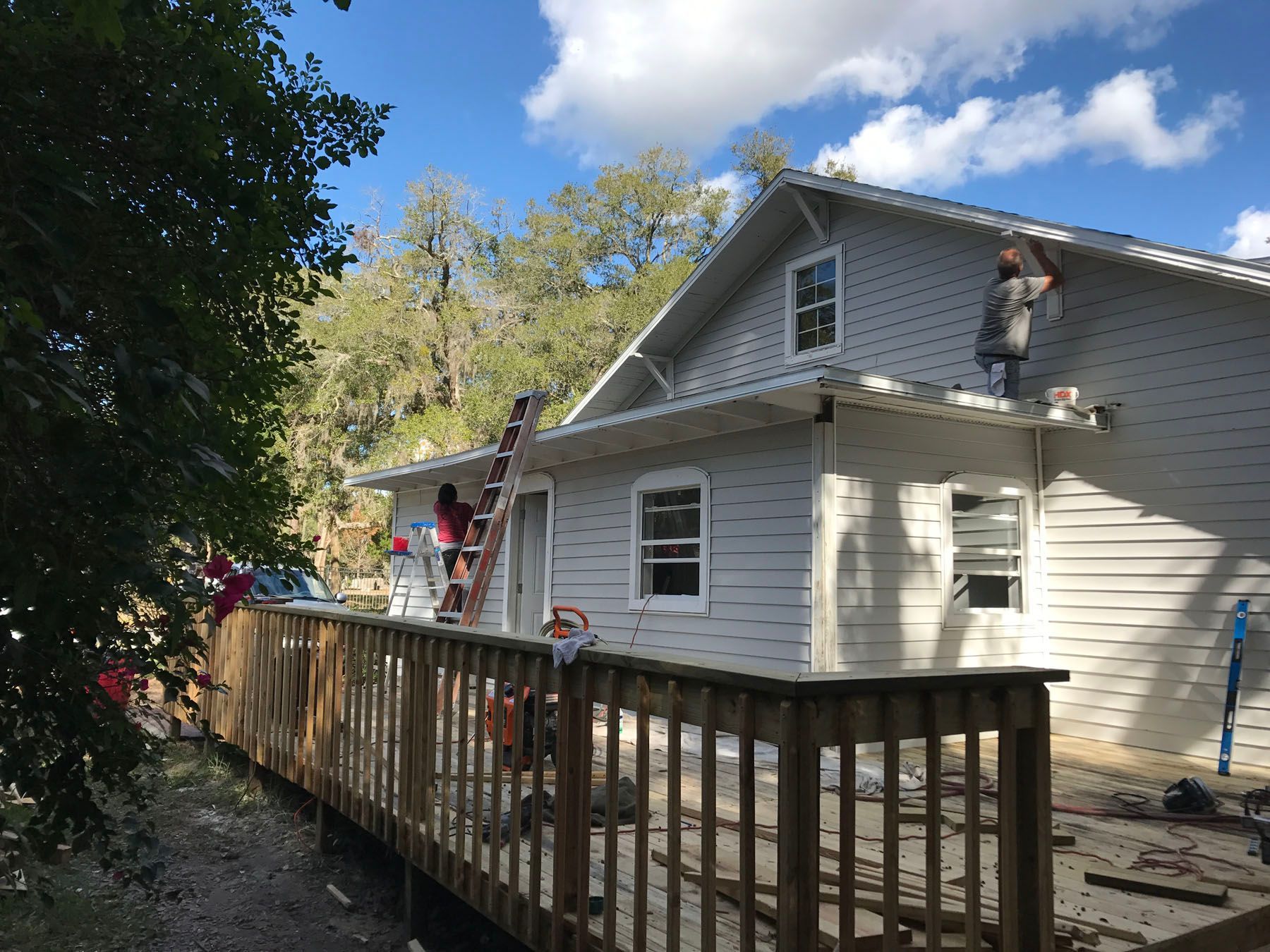 Two people work on a house exterior; one stands on a ladder and the other on the roof, painting light-colored siding.
