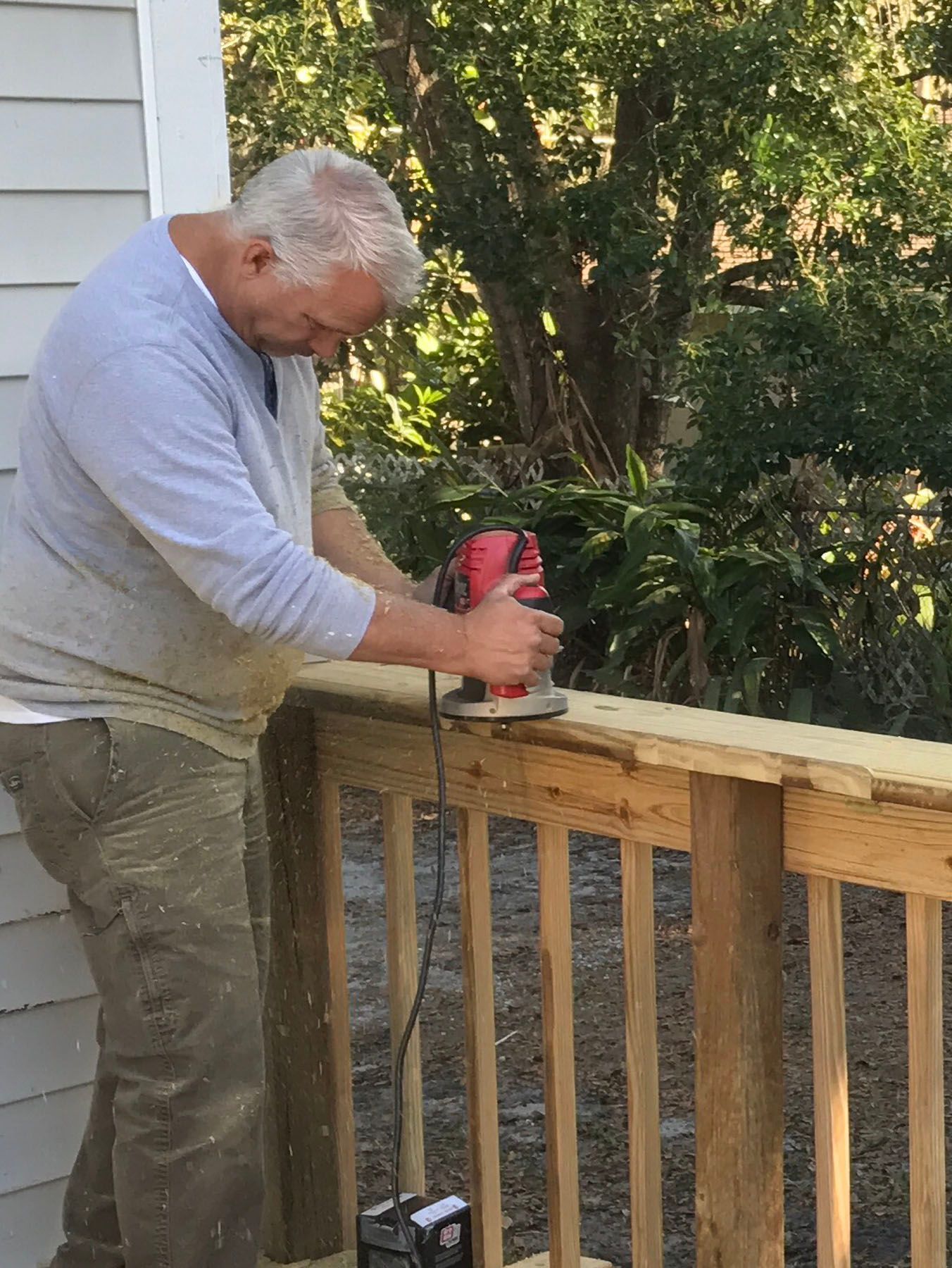 A person uses a red power router to smooth the edge of a wooden railing on an outdoor deck.
