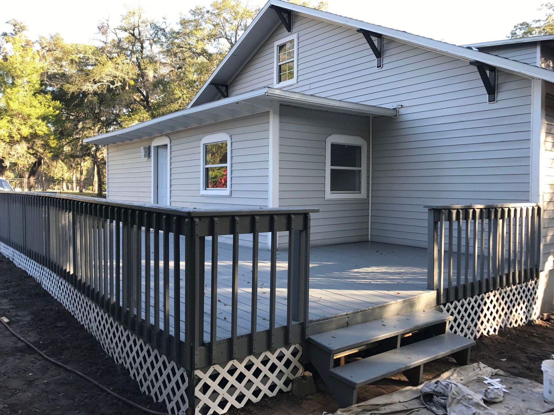 Light grey house with a large, dark grey wooden deck, steps, and white lattice skirting in a sunny, wooded area.