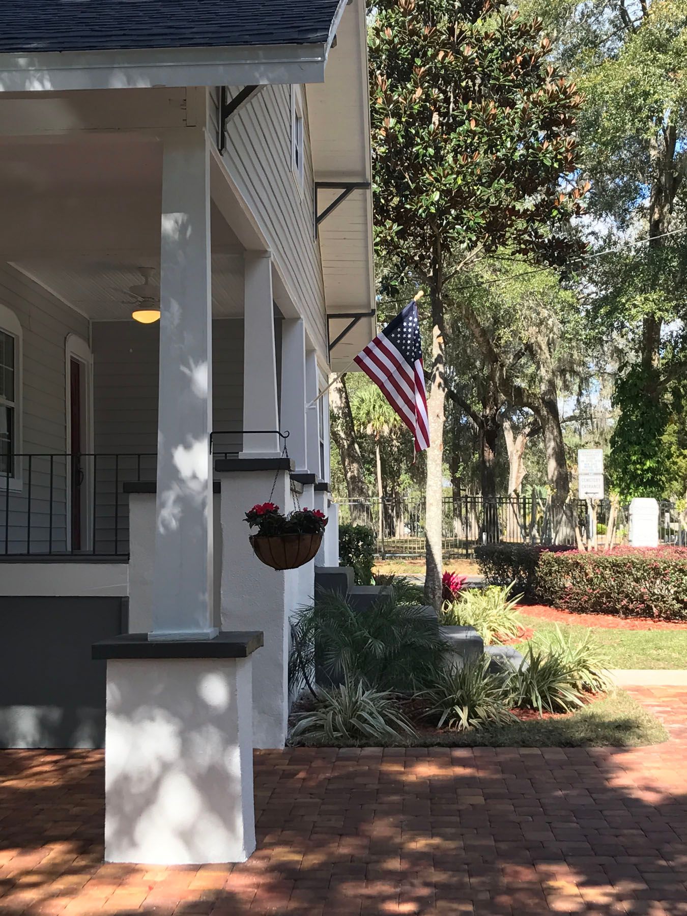 A white porch with brick pavers, a hanging flower basket, and an American flag visible in the background trees.