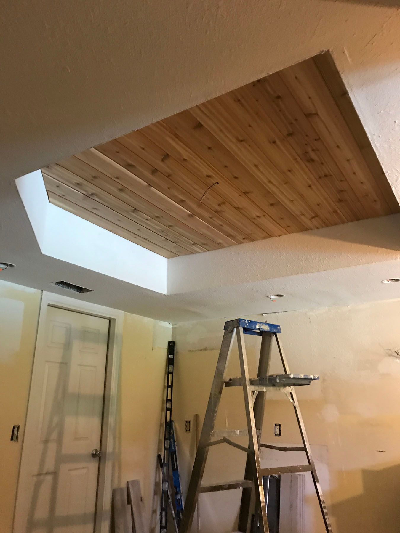 A room under renovation featuring a recessed ceiling with wooden paneling, a ladder, and unfinished wall surfaces.