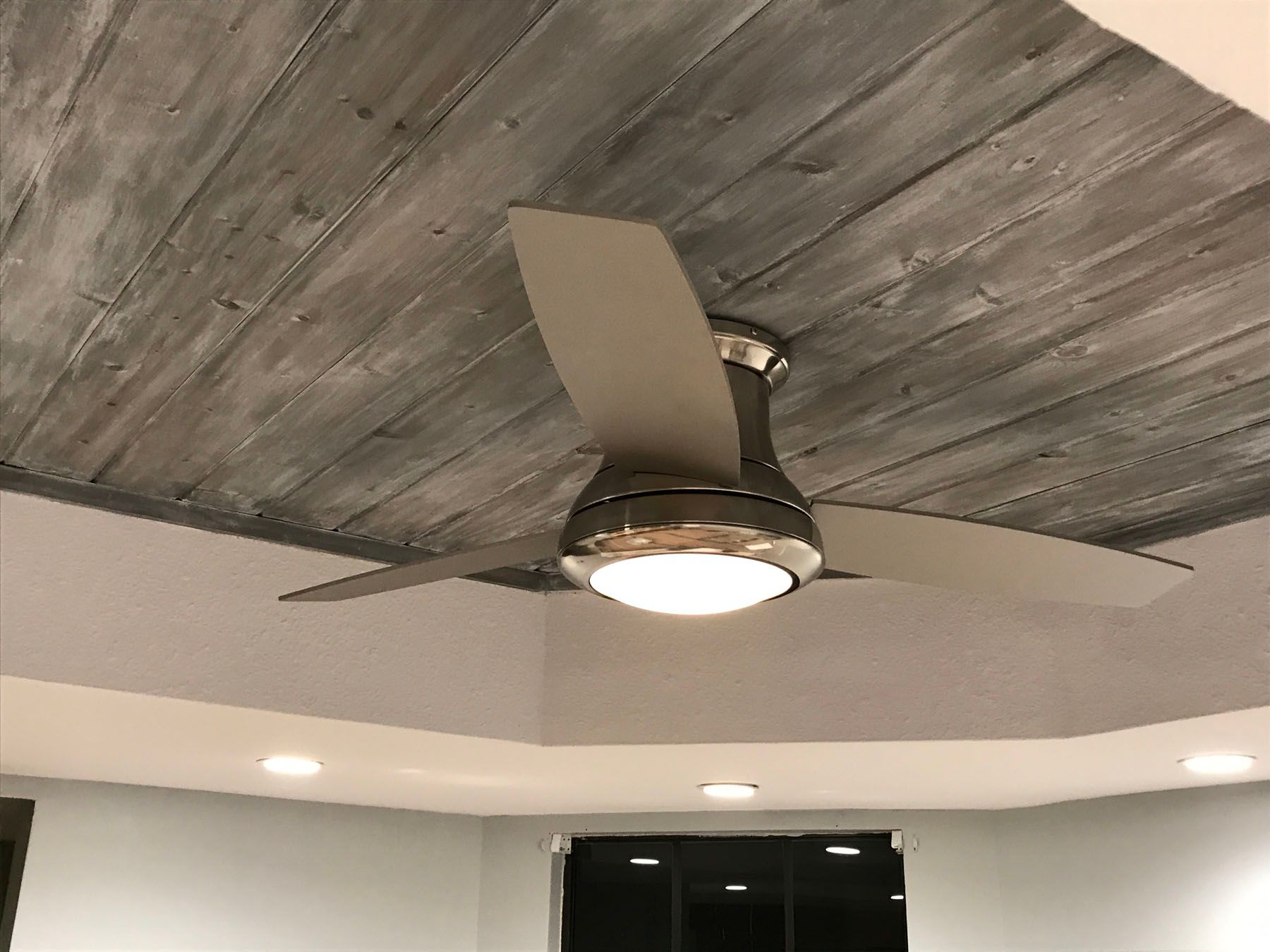 A ceiling fan with a built-in light mounted to a grey, wood-paneled tray ceiling in a room.