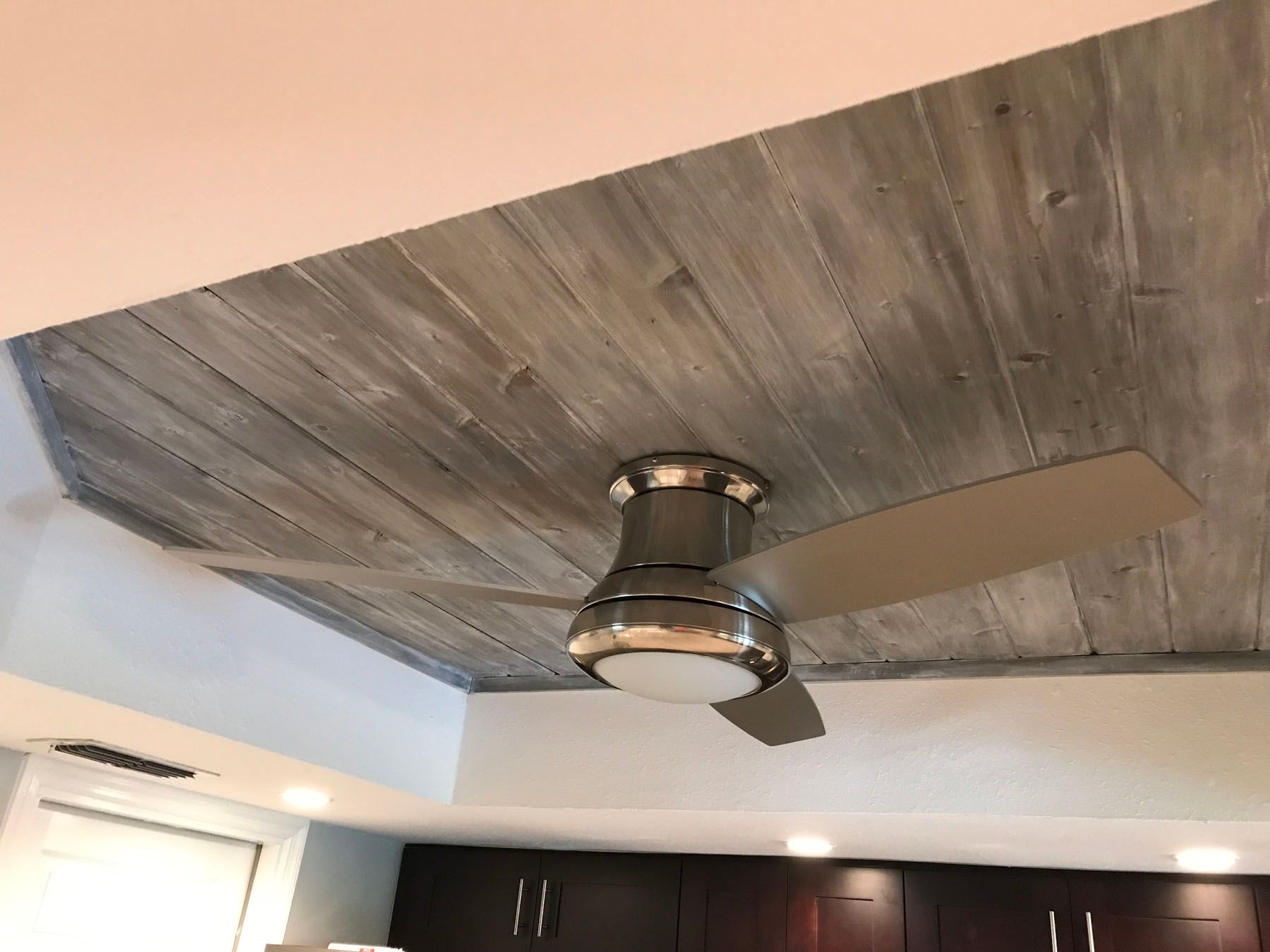 A ceiling fan with a built-in light mounted on a wooden-planked ceiling in a room with white tray ceiling accents.