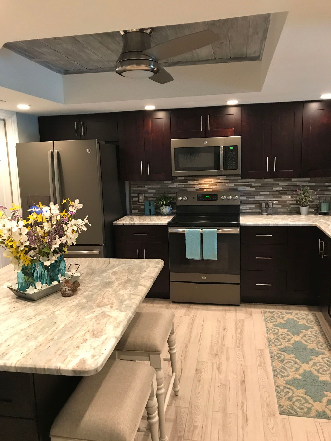 A modern kitchen featuring dark cabinets, light granite countertops, an island with two stools, and a textured ceiling.