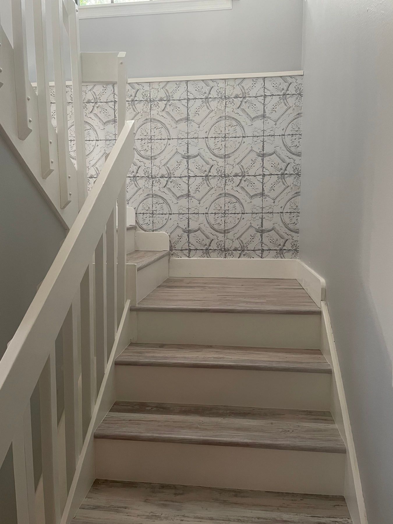 A wooden staircase with white railings, featuring gray patterned wallpaper on the adjacent wall.