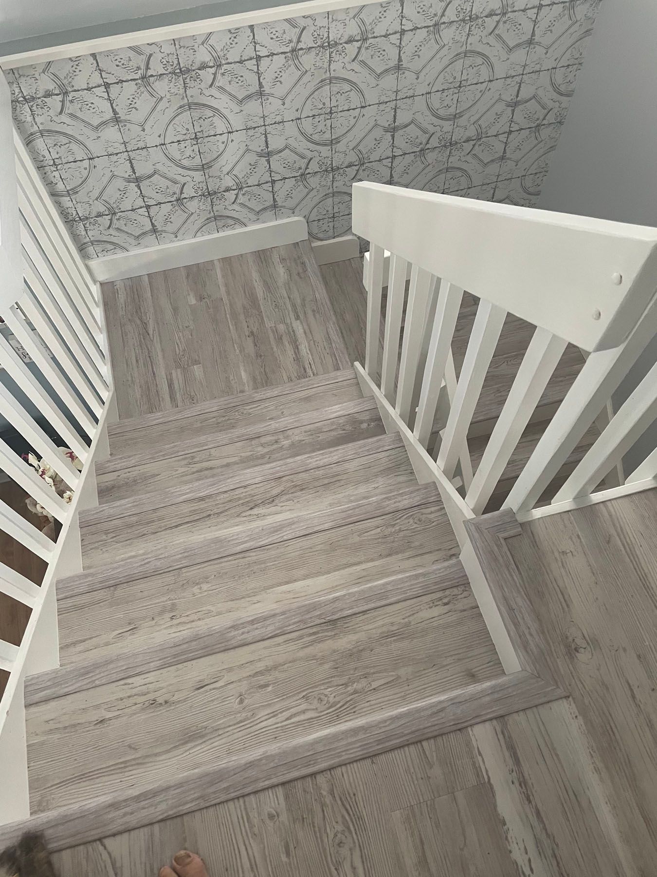 Top-down view of gray wood-look stairs with a white railing and patterned wallpaper on the landing wall.