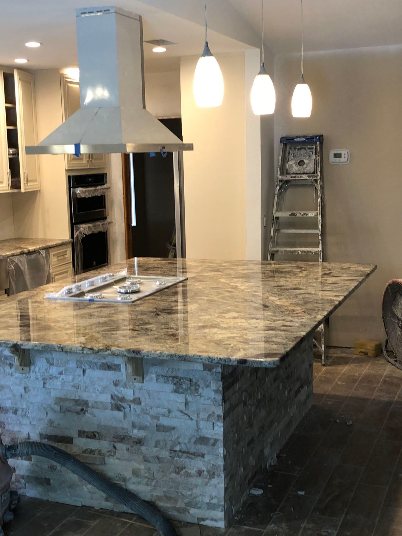 A kitchen island with a stone base and granite countertop, featuring a stovetop, overhead hood, and three pendant lights.