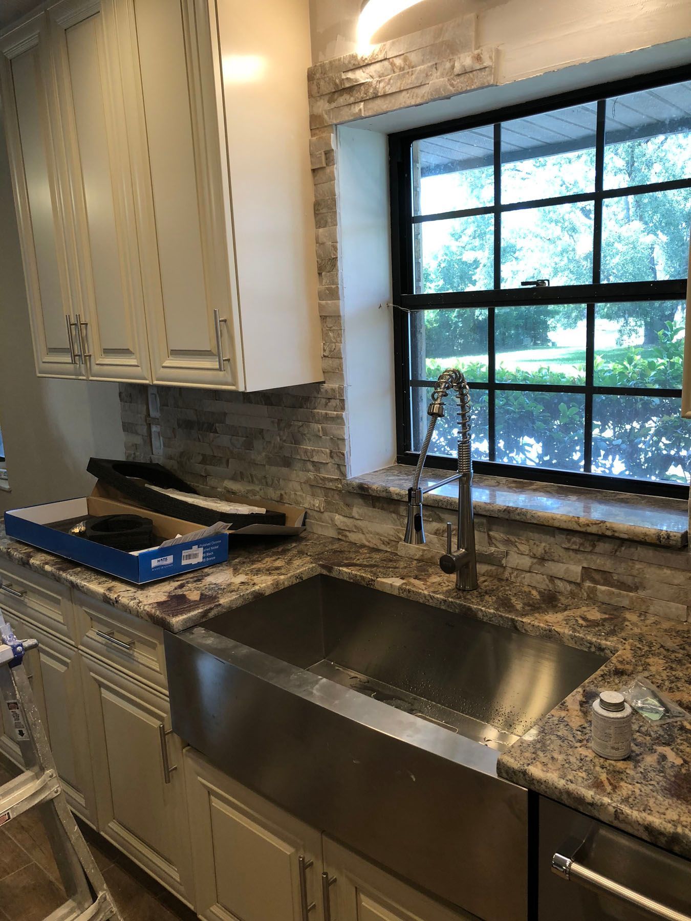 A kitchen sink area featuring a stainless steel farmhouse sink, granite countertops, and stone backsplash under a window.
