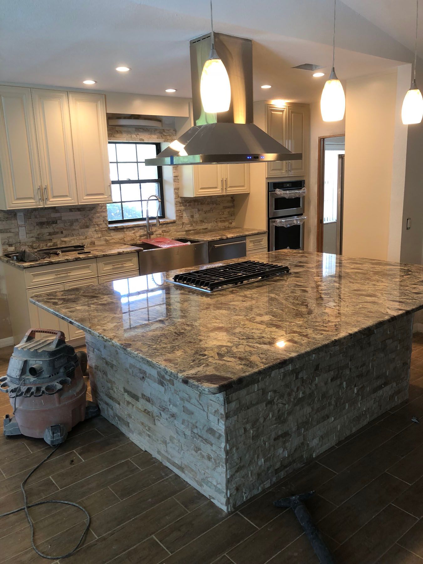 A renovated kitchen featuring a large stone-base island with a granite countertop, gas cooktop, and hanging pendant lights.