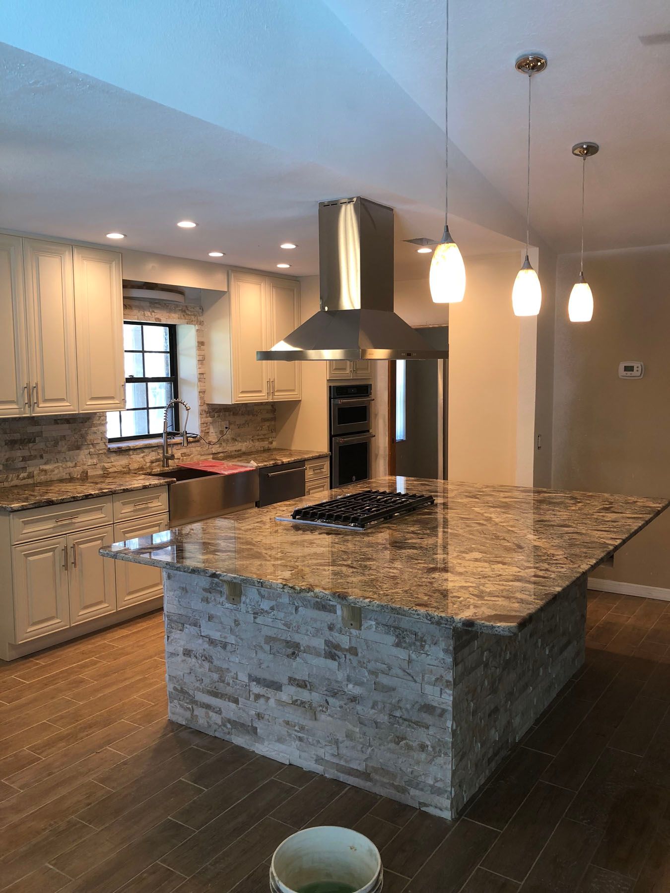A kitchen features a large stone-base island with a cooktop, granite countertops, light cabinets, and three pendant lights.