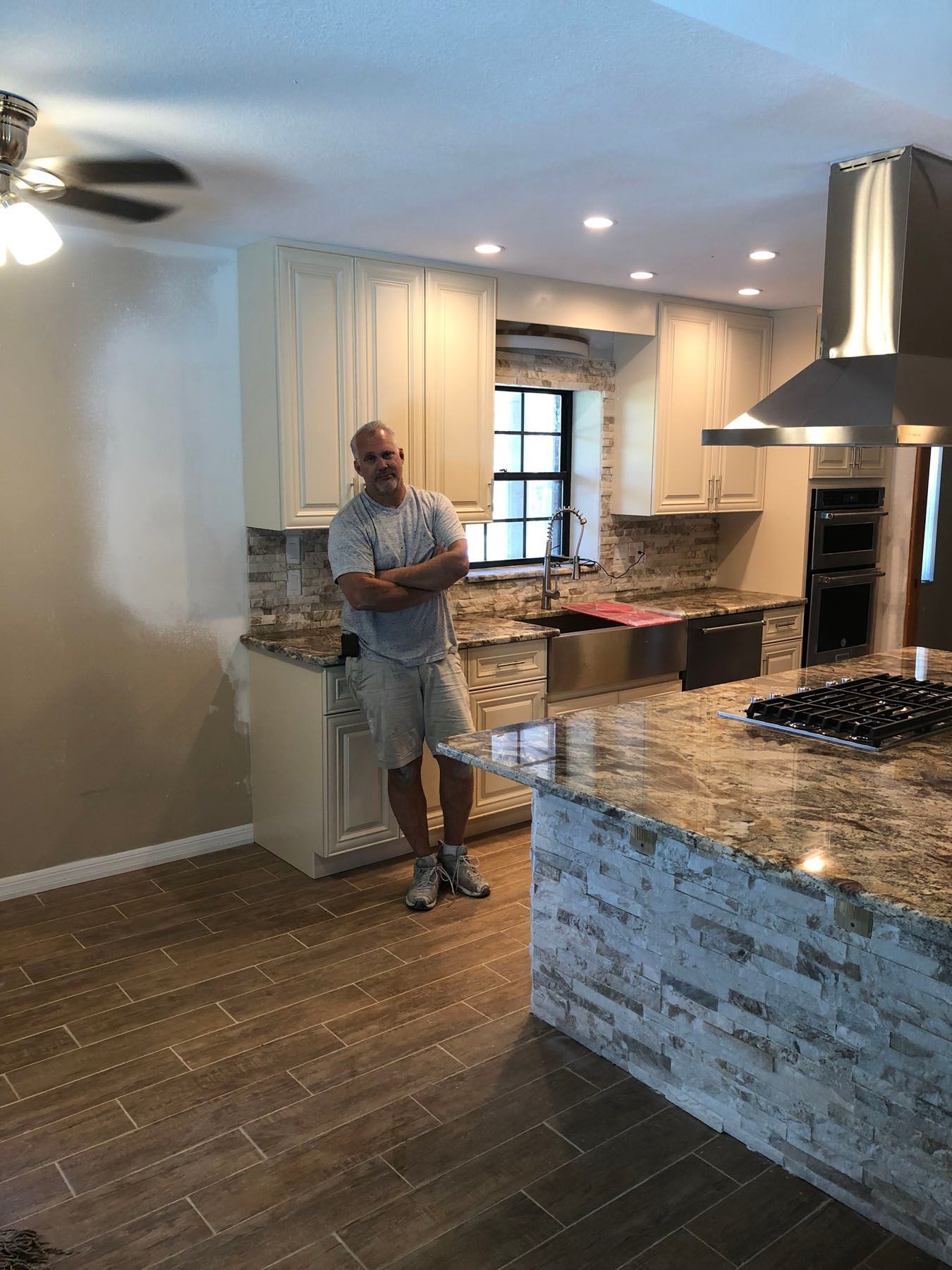 A man stands with arms crossed in a modern kitchen with white cabinets, granite countertops, and wood-look tile floors.