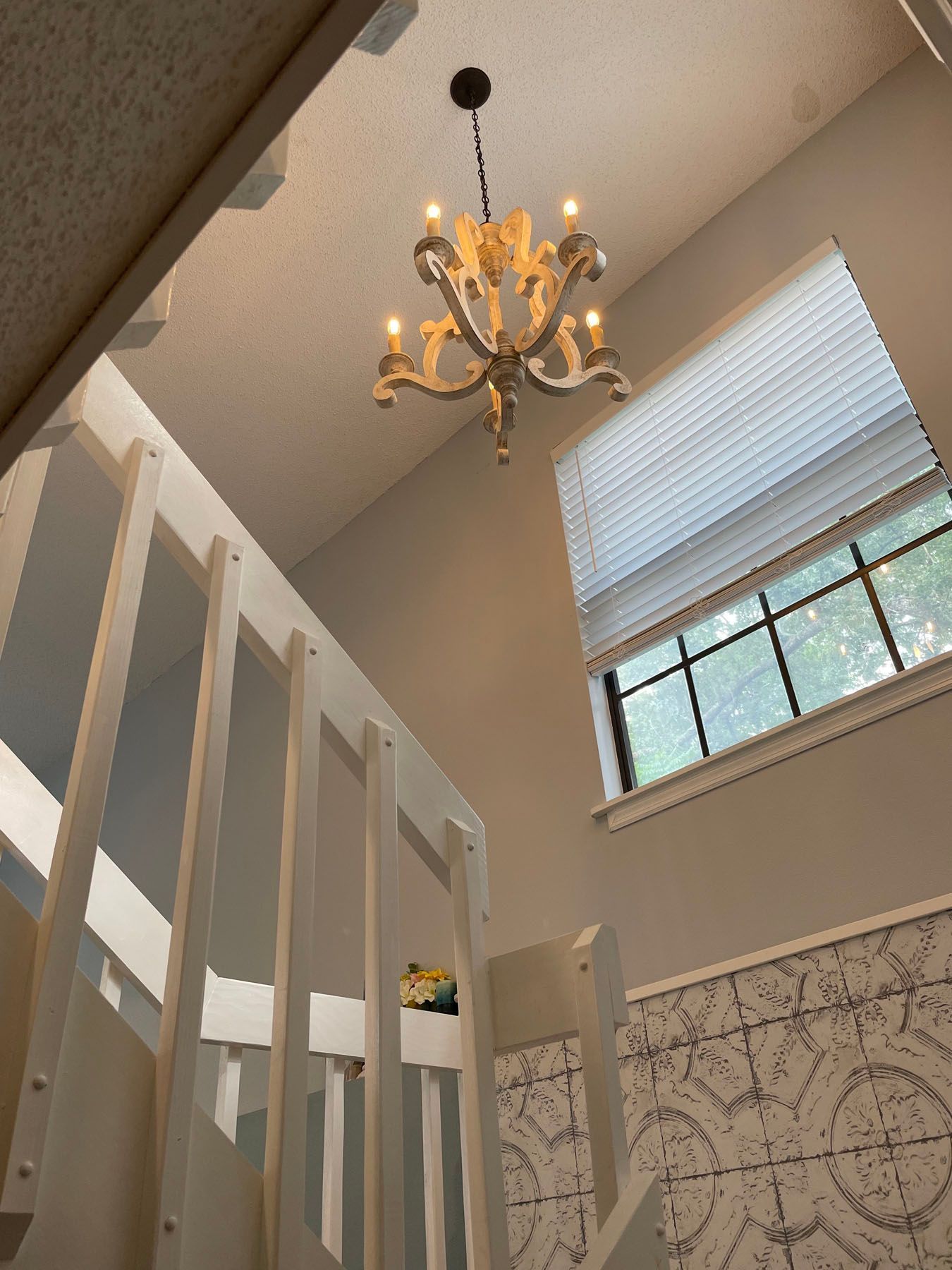 A staircase with white wooden railings leads up toward a lit chandelier hanging in front of a window with blinds.