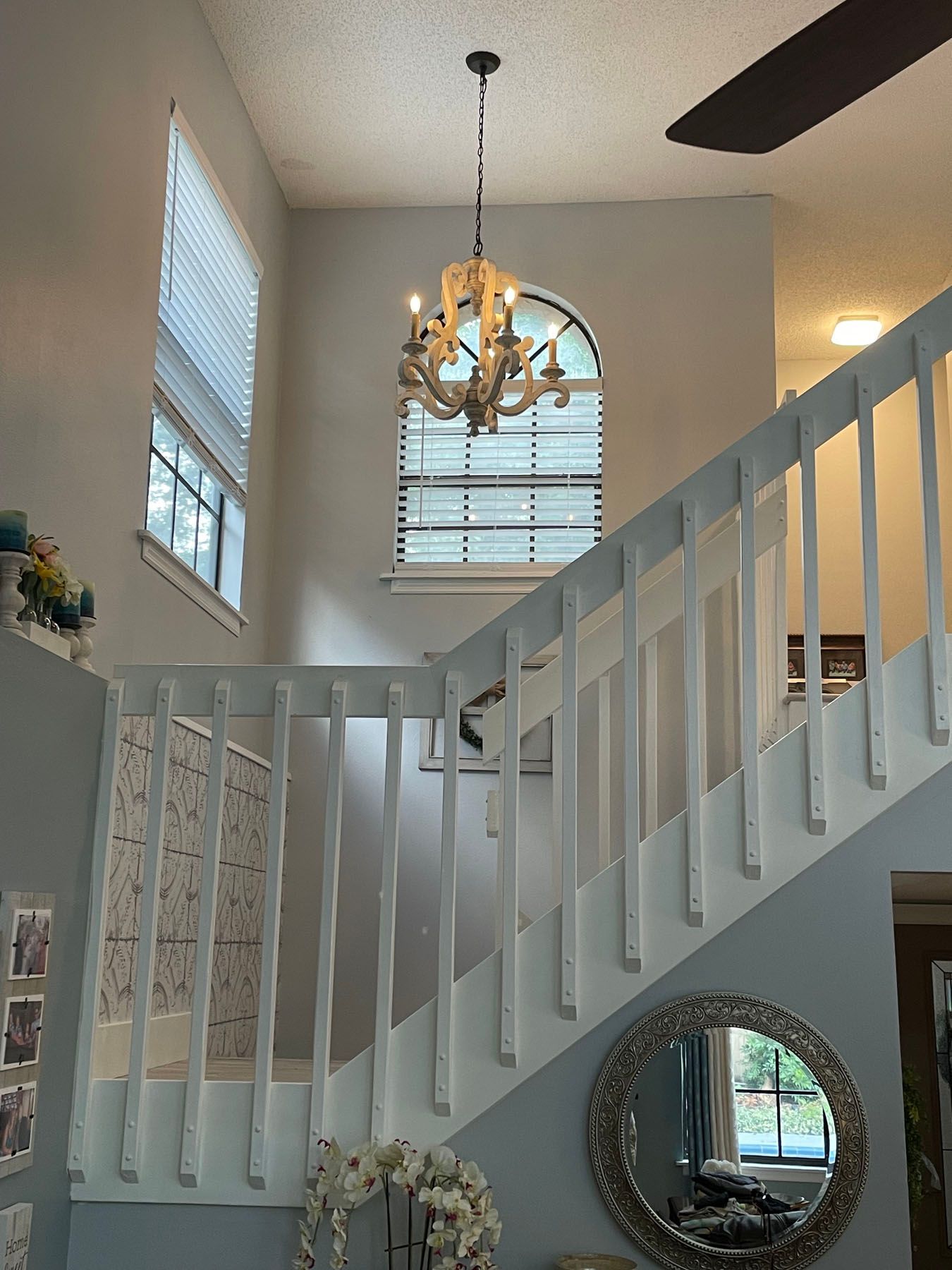A white wooden staircase with a chandelier hanging in front of a large arched window in a light-colored home foyer.