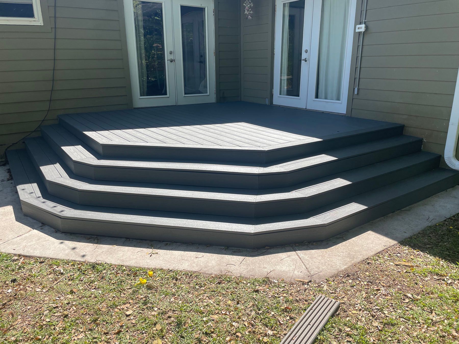 A three-tiered, grey, hexagonal wooden deck leading to two white sets of French doors on the side of a house.
