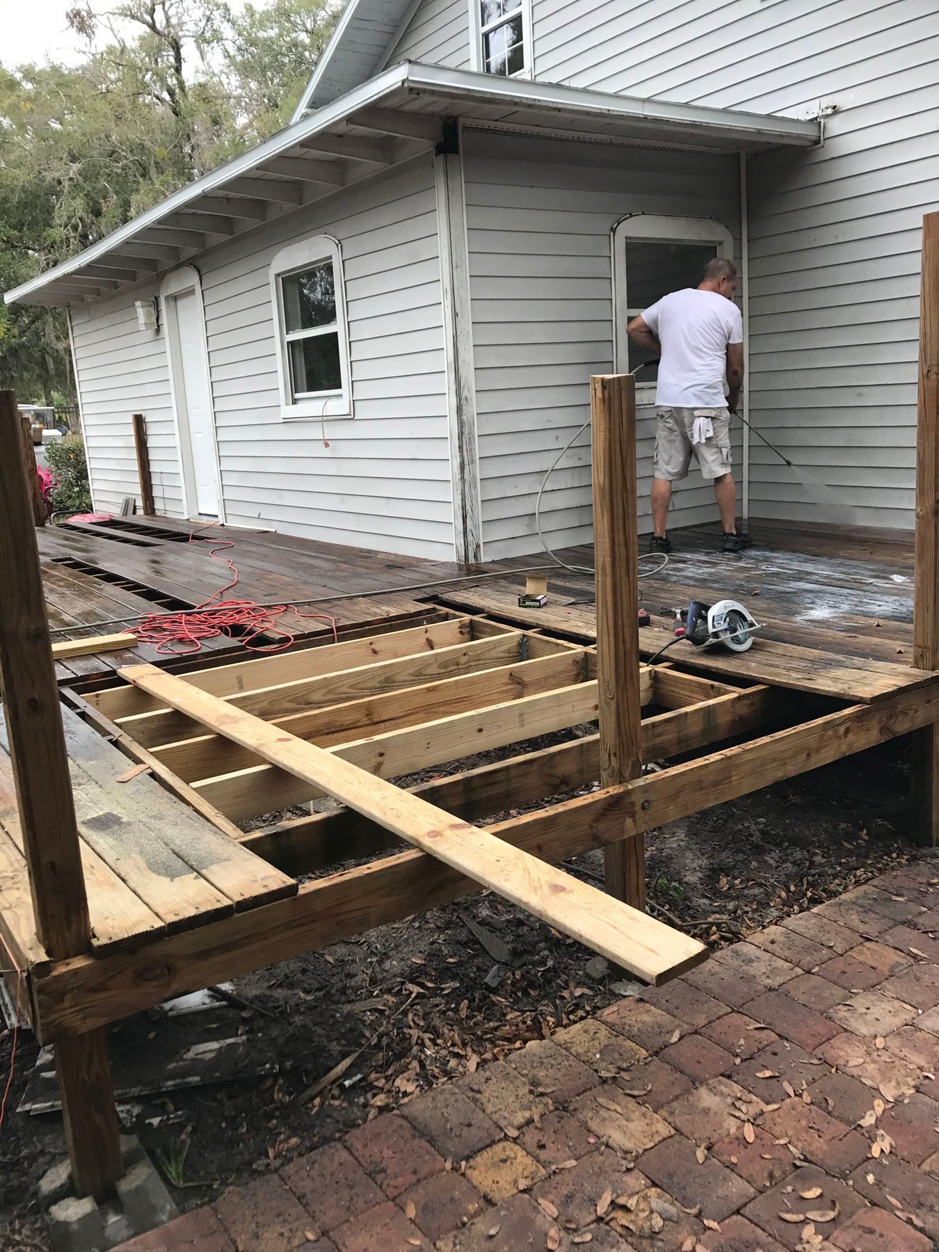 A person in a white shirt and shorts works on a wooden deck frame attached to the side of a light gray house.