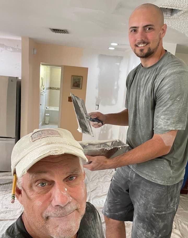 Two people performing drywall repairs in a home renovation setting, one holding a taping knife and mud tray.