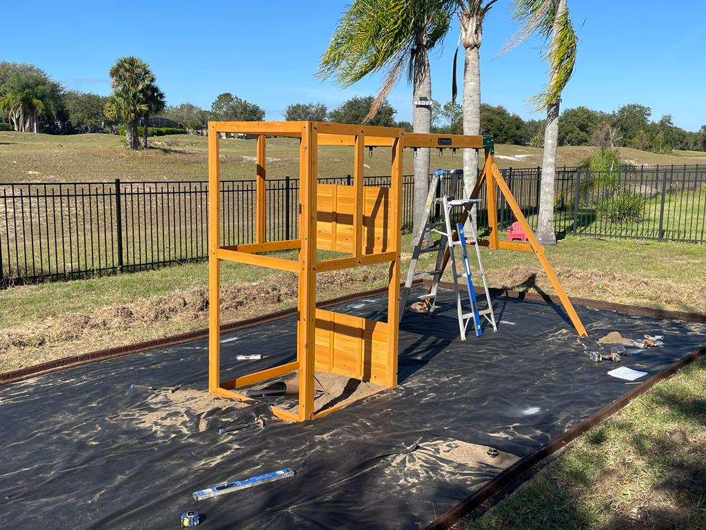 A partially assembled wooden playset sits on a black landscape fabric-covered area in a yard with trees and a fence.