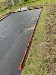 An orange leveling tool resting on a metal border strip over a black landscape fabric patch in a grassy yard.