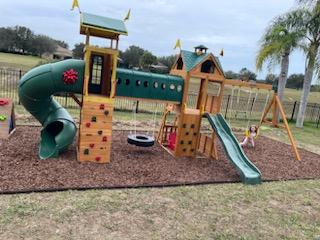 A large wooden outdoor playground set with a green tube slide, climbing wall, tire swing, and small slide on wood mulch.