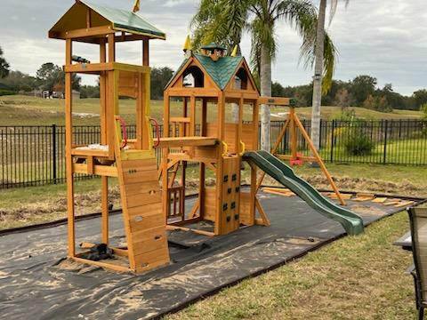A wooden backyard playset with two towers, a green slide, and a swing set, installed on a landscape fabric base.