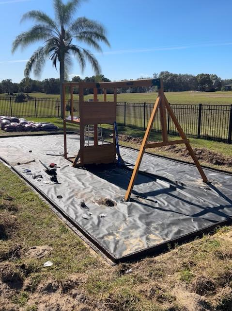 Wooden swing set frame under construction on a landscape fabric base in a backyard with a palm tree and fence.