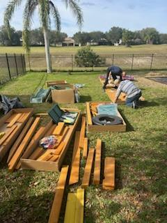 Two people assemble wooden garden beds in a grassy yard, surrounded by scattered lumber, tools, and cardboard packaging.