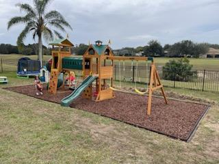 A wooden playground set with a green roof, slide, and swings, situated on a mulch bed in a grassy backyard.