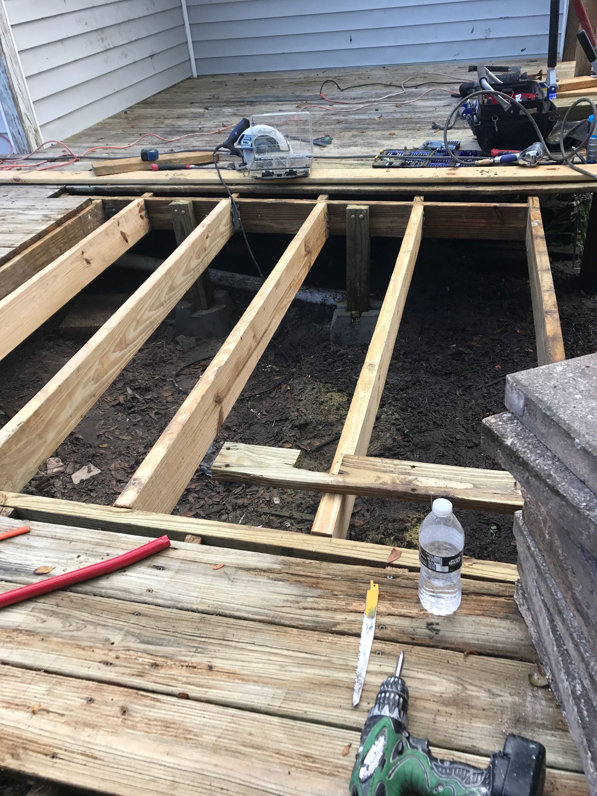 A deck under construction with exposed wooden joists, scattered tools, a water bottle, and a stack of pavers.