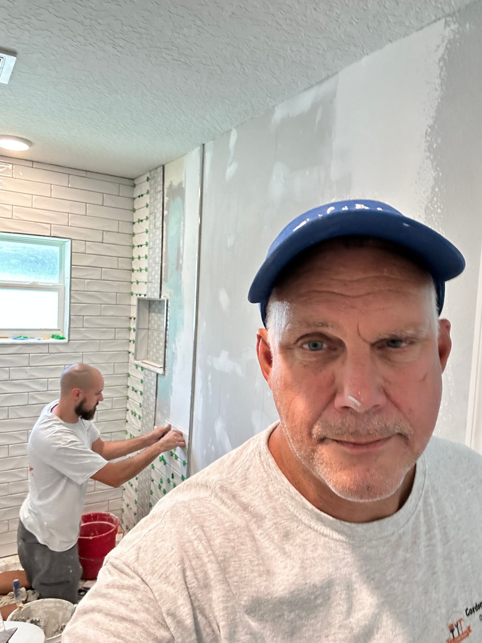 A man in a blue cap takes a selfie while another person works on a tiled wall in the background of a room under renovation.