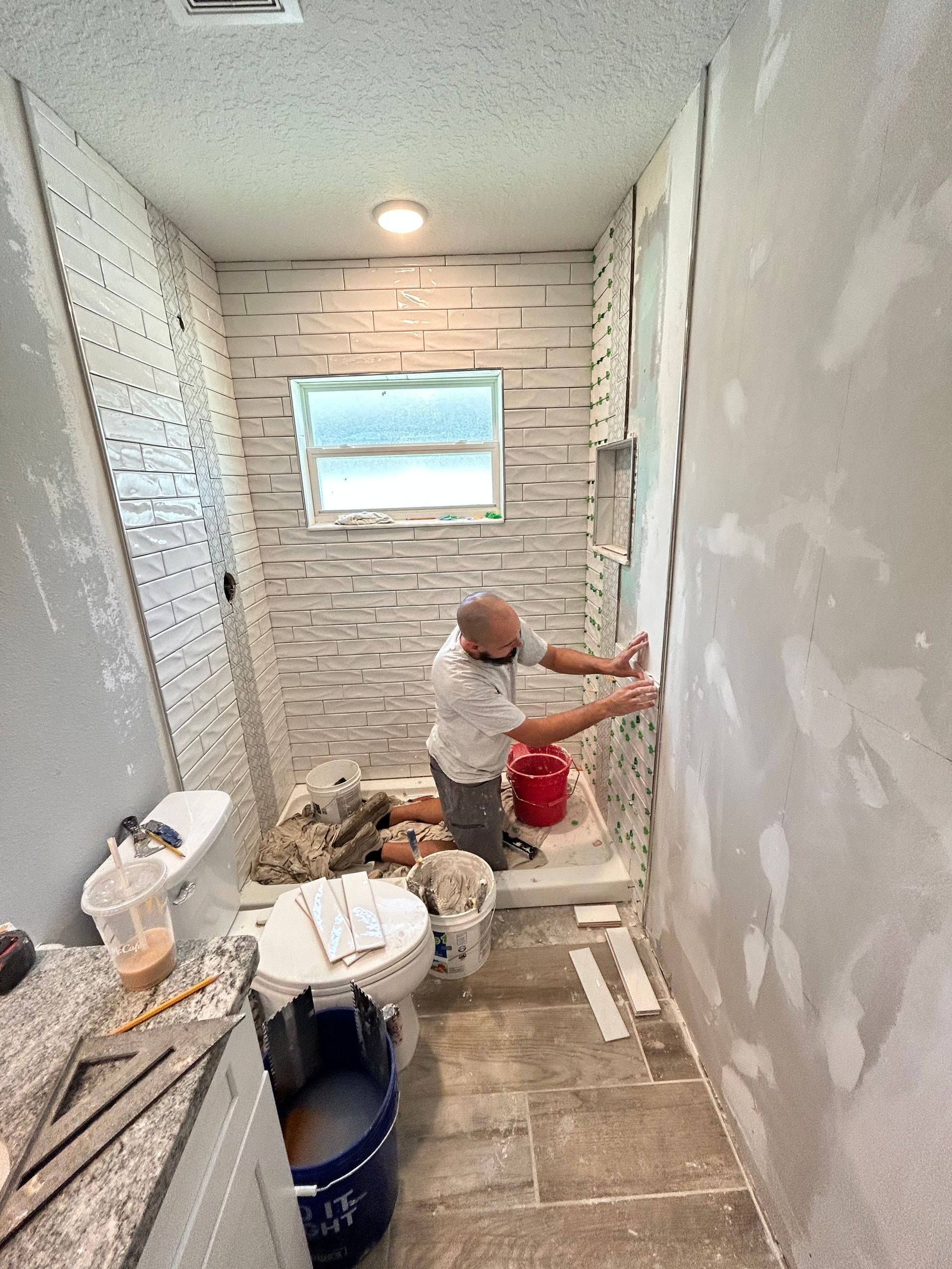 A worker installs white, textured stone tiles in a bathroom shower area with unfinished walls and a tile floor.