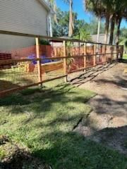 A wooden rail fence with wire mesh lines a yard next to a tan house under a clear blue sky.
