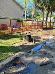 A wooden post-and-rail fence stands in a grassy yard, with a dark trash can and a narrow, muddy drainage ditch in the front.
